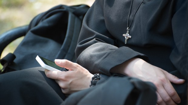 Unrecognizable-nun-in-a-black-robe-with-a-cross-on-his-neck-uses-a-smartphone-